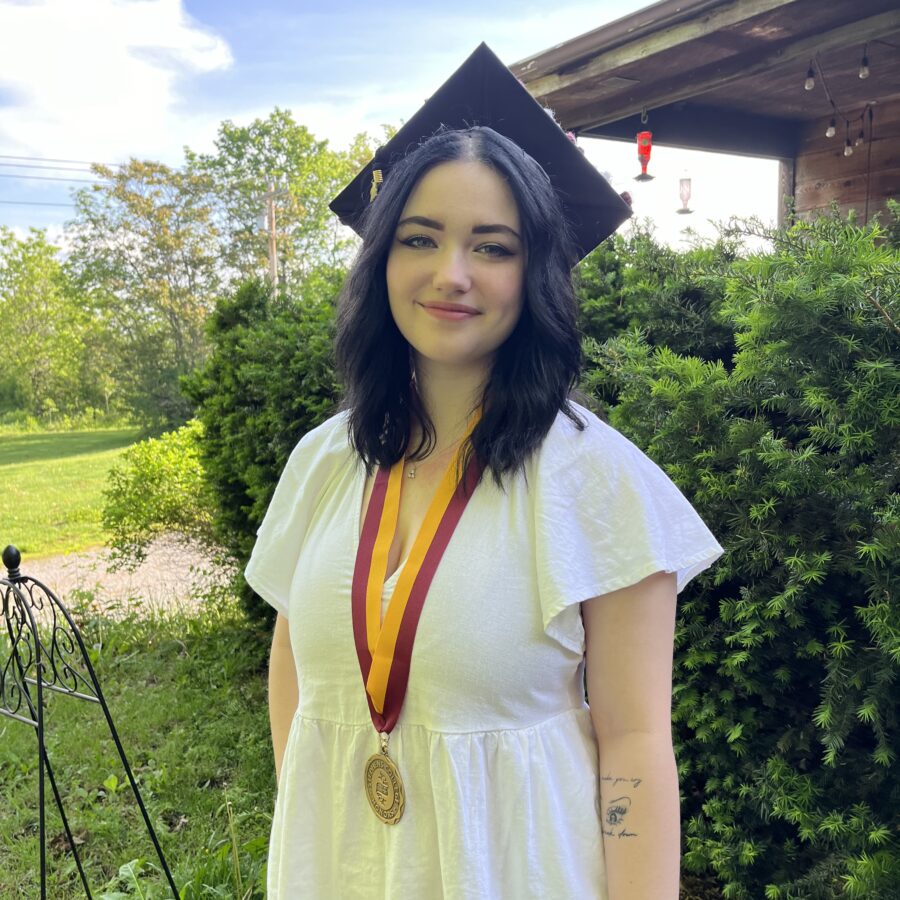 A graduate smiles outside of a house on a sunny day. She's wearing a white dress, a graduation cap and an honors medal.