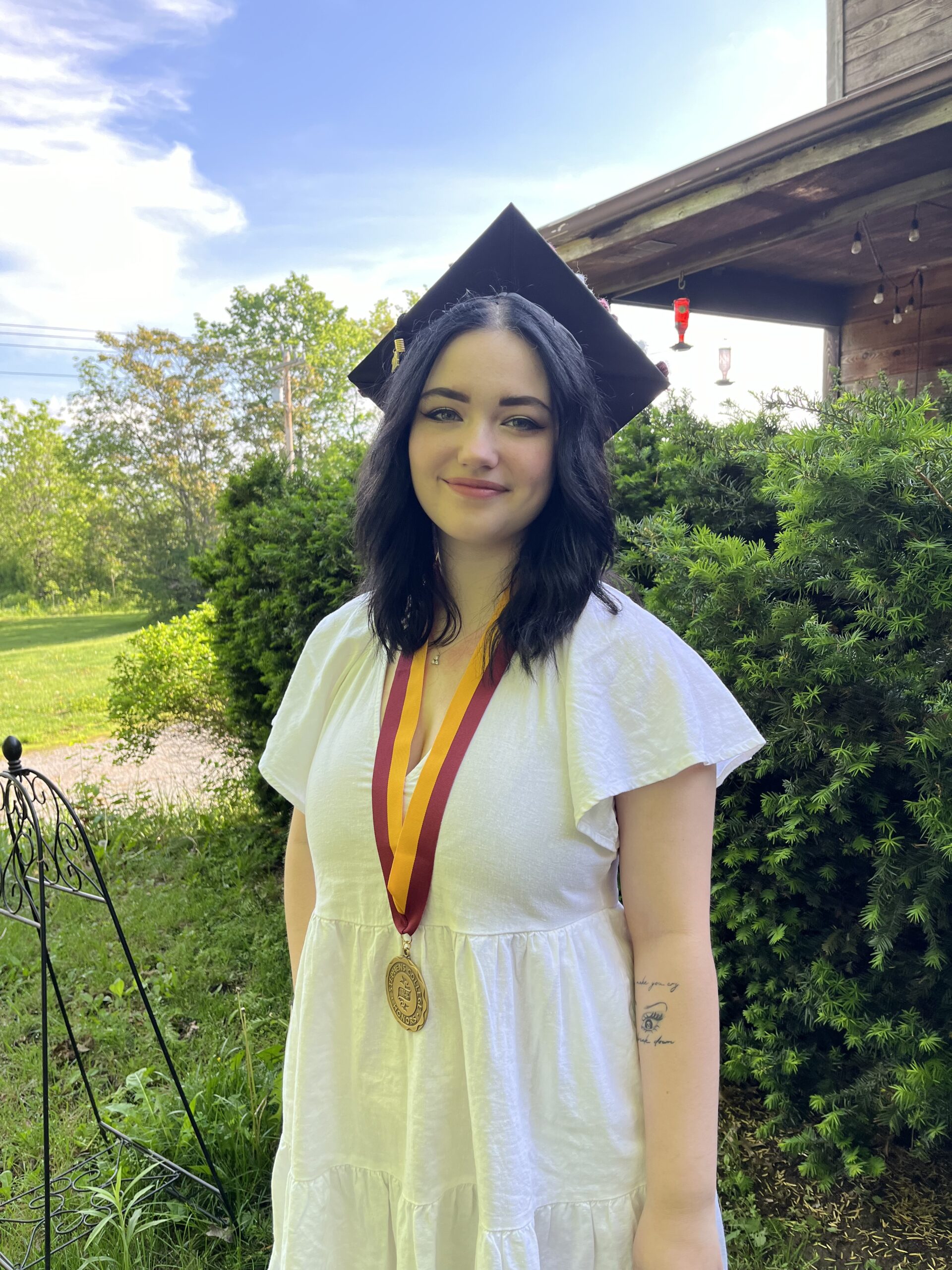 A graduate smiles outside of a house on a sunny day. She's wearing a white dress, a graduation cap and an honors medal.