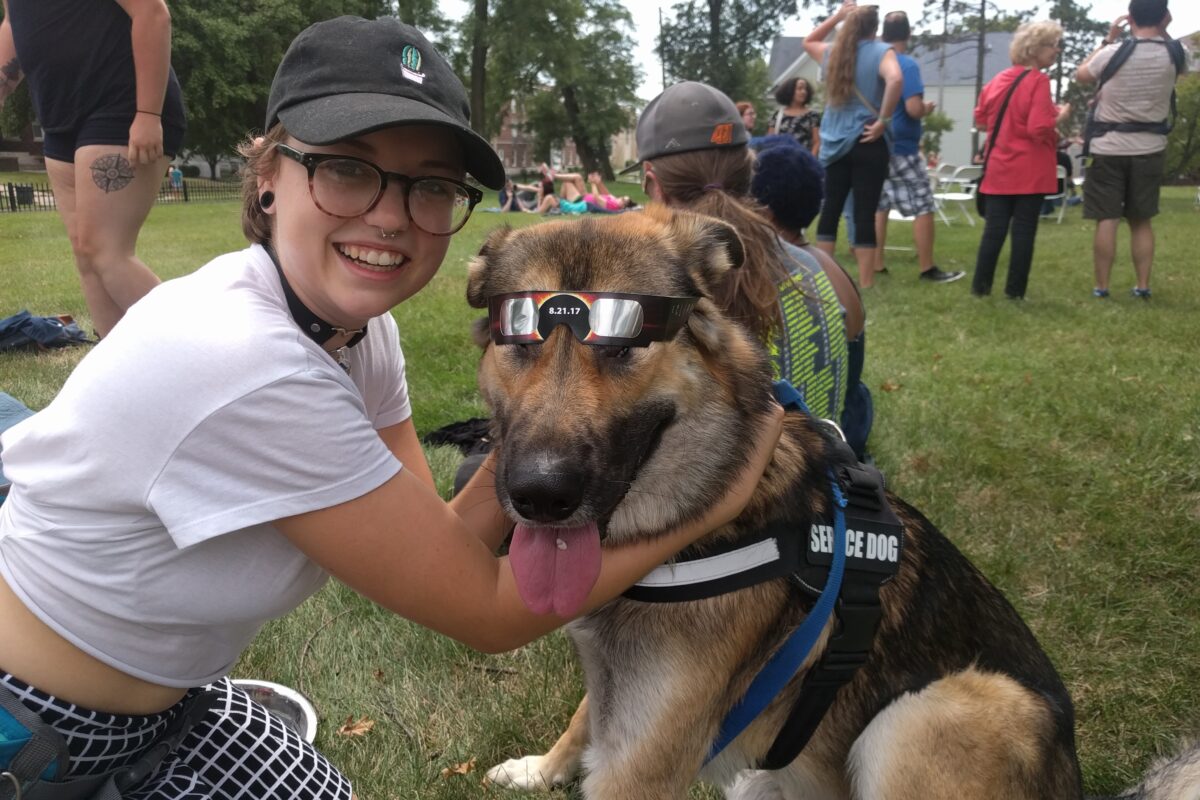 A student poses with her assistance dog, who is wearing protective eyewear for viewing an eclipse.