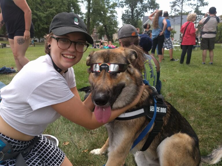 A student poses with her assistance dog, who is wearing protective eyewear for viewing an eclipse.