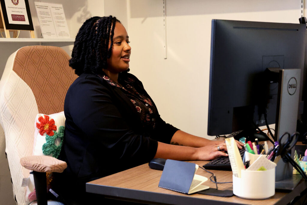 Shana Nunn works at a desk with a large computer monitor. Office supplies, a notebook, and a pen holder sit on the desk. A decorative pillow rests on the chair, and certificates and books are displayed on a wall shelf.