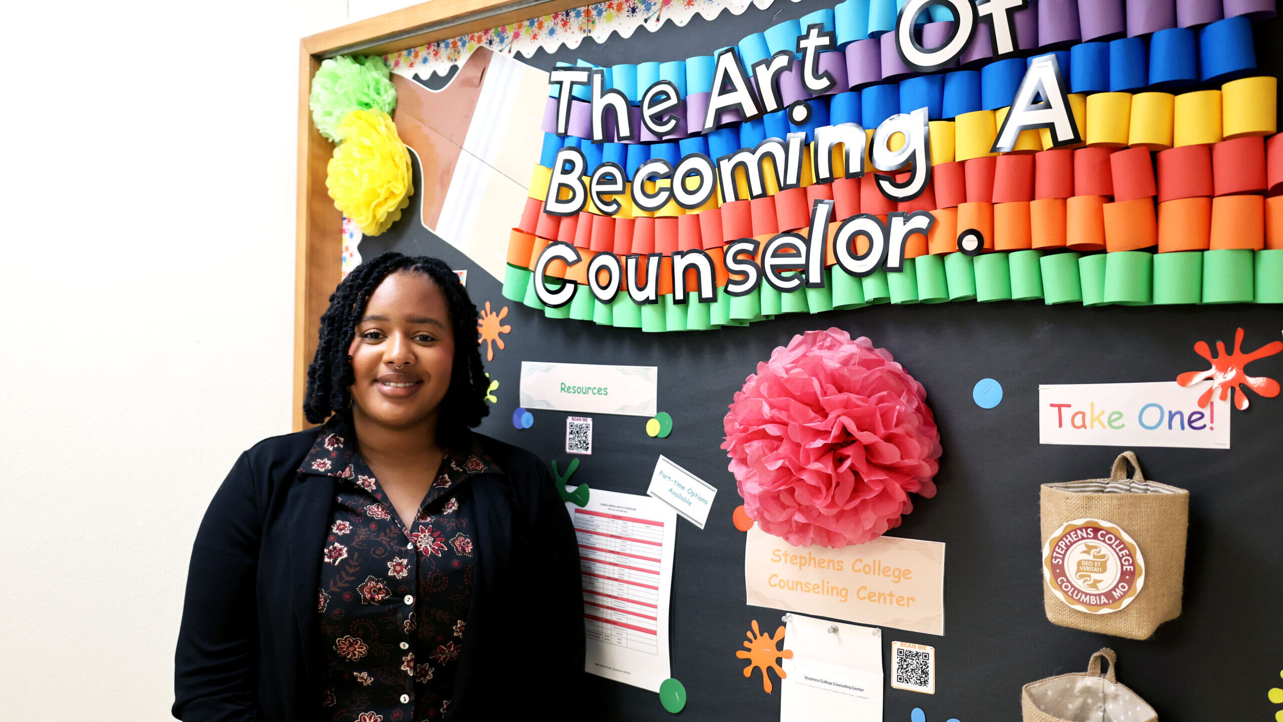 Shana Nunn stands beside a bright, multicolored bulletin board titled “The Art of Becoming a Counselor.” The board includes paper decorations, counseling center information, and resource handouts.