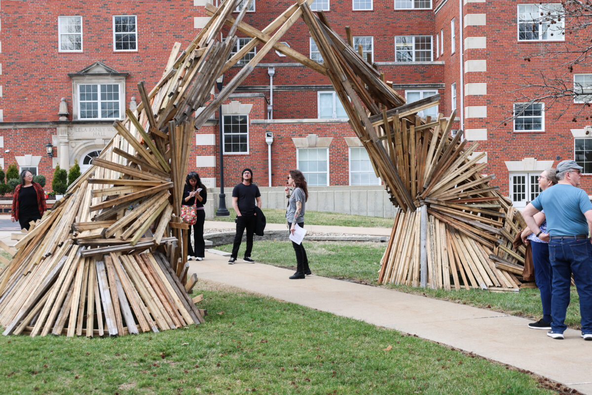 A large wooden outdoor sculpture made of stacked and angled wooden planks arranged in two leaning towers that form an arch over a sidewalk. People stand around the sculpture, walking through and observing it. Brick residential buildings fill the background.