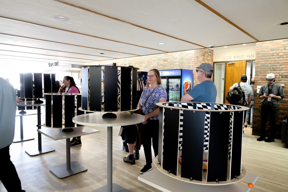 A lobby area with several cylindrical black-and-white optical illusion devices arranged on round tables. People stand around the displays, observing and rotating them. In the background, there is a vending area, brick walls, and a hallway entrance.