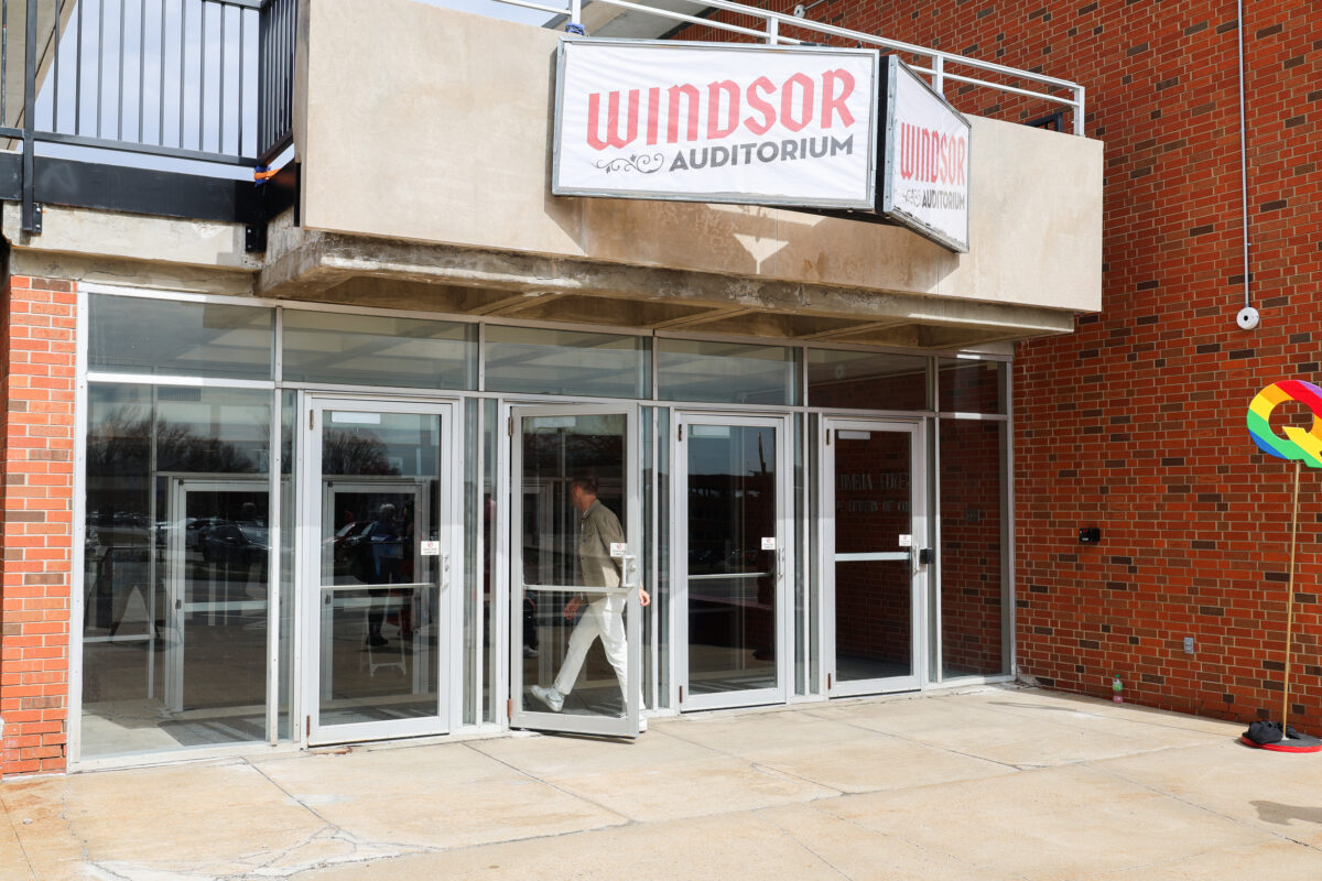 Exterior view of the Windsor Auditorium entrance. Large glass doors line the front of the building, and a sign reading “Windsor Auditorium” hangs above the doorway. A person is entering through one of the center doors. The building has brick walls and a concrete overhang.
