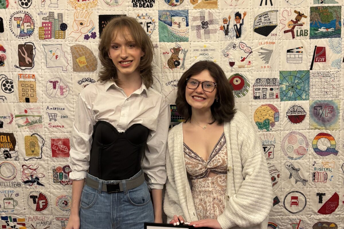 Two people stand in front of a large quilt-like backdrop made of many individually designed square fabric panels featuring drawings, symbols, and text. In front of them is a small sign with event information on a stand. The photo is taken indoors.