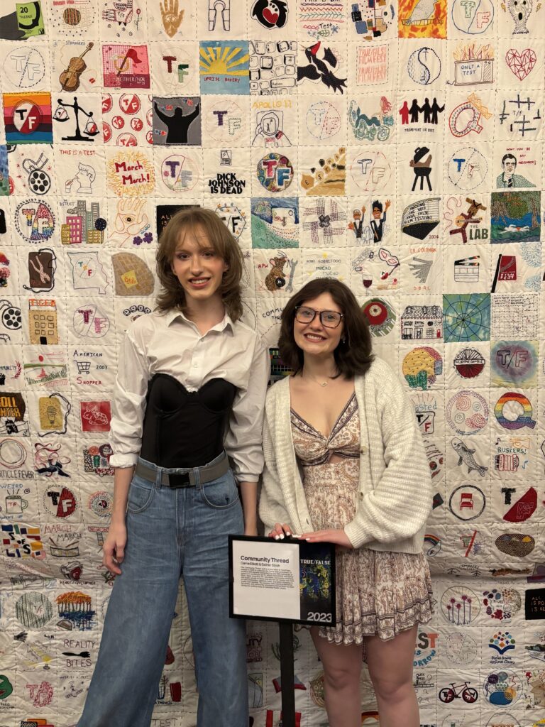 Two people stand in front of a large quilt-like backdrop made of many individually designed square fabric panels featuring drawings, symbols, and text. In front of them is a small sign with event information on a stand. The photo is taken indoors.